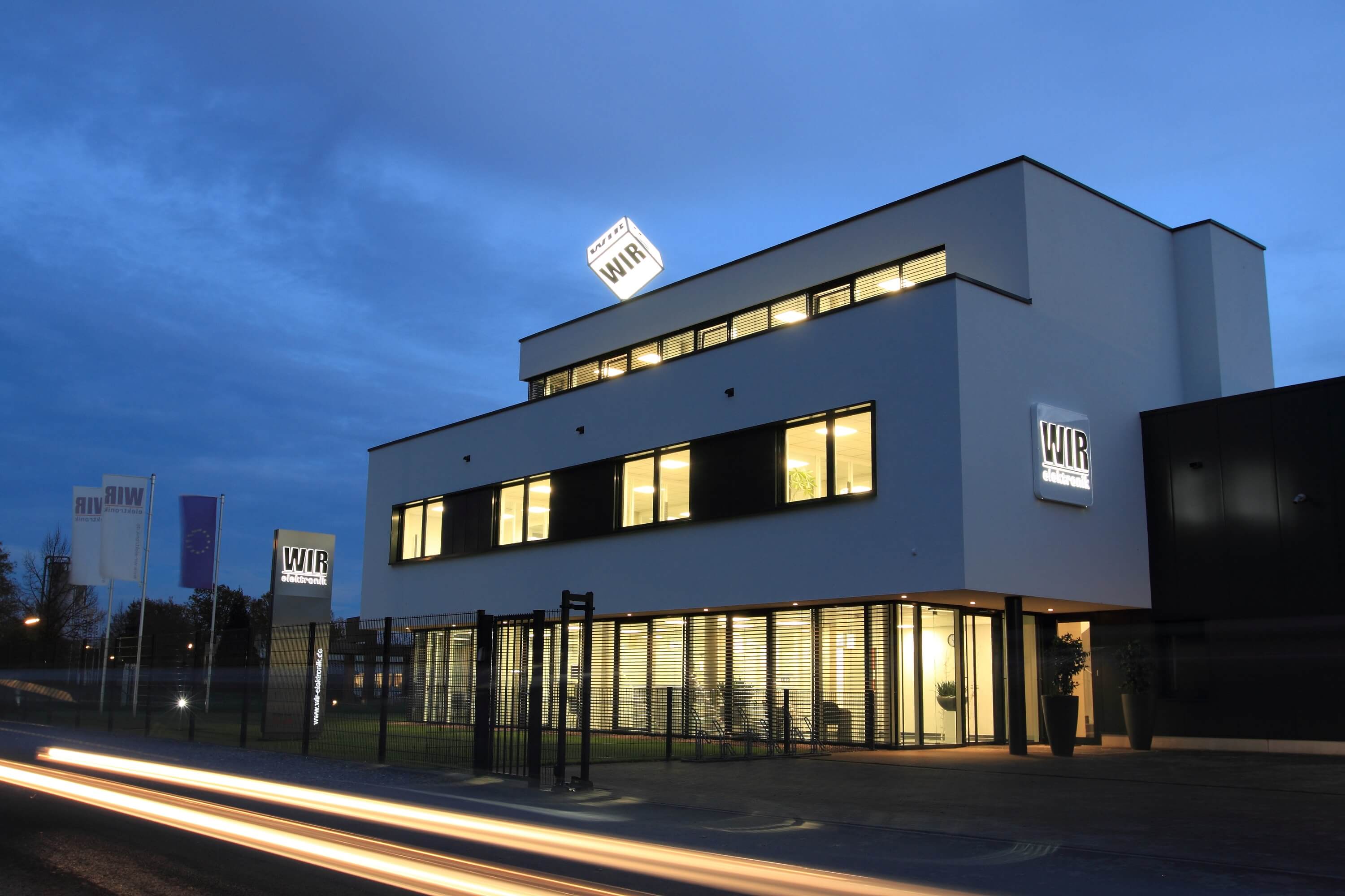 Verwaltungsgebäude_Wir-Elektronik_beleuchtet_Dunkelheit_Logos_Fahnen